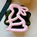 Minimalist pink bunny-shaped silicone baby teether placed on a textured beige surface next to a green ribbon with gold accents.