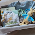 A premium baby gift box featuring a blue cotton bodysuit, a Peter Rabbit plush toy, a "Guess How Much I Love You" soft book, and a sage green elephant teether, all laid out on white tissue paper in a cardboard mailer box.