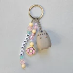 A cute gray Pusheen Tropical Vacation Keyring with pastel pink, yellow, and blue letter beads spelling PUSHEEN, attached to a durable silver ring.